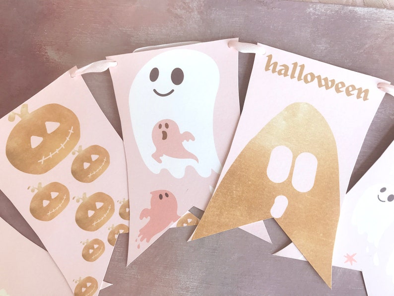 May include: A decorative banner featuring light pink pennants strung on a ribbon. The pennants display gold foil jack-o'-lanterns, white ghosts with smiling faces, smaller pink ghosts, and a gold foil ghost shape with the text halloween.