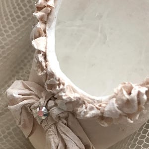 May include: Close-up of a decorative, off-white, oval-shaped object with a ruffled edge. A fabric bow with a crystal accent adorns the side. The object has a soft, textured appearance, suggesting a delicate craft or design.