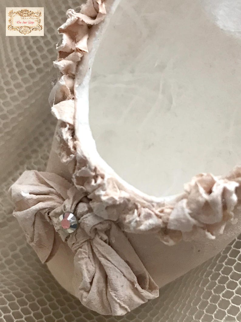 May include: Close-up of a decorative, off-white, oval-shaped object with a ruffled edge. A fabric bow with a crystal accent adorns the side. The object has a soft, textured appearance, suggesting a delicate craft or design.