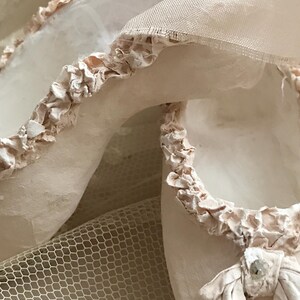 May include: Close-up of a pair of off-white ballet slippers with delicate, ruffled trim. The slippers are made of soft fabric and feature a small bow with a decorative jewel. A fine mesh fabric is visible in the background.