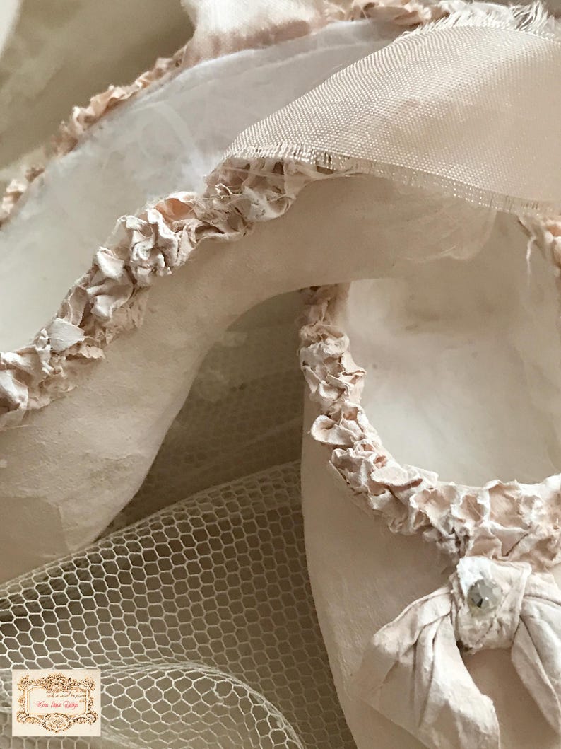 May include: Close-up of a pair of off-white ballet slippers with delicate, ruffled trim. The slippers are made of soft fabric and feature a small bow with a decorative jewel. A fine mesh fabric is visible in the background.