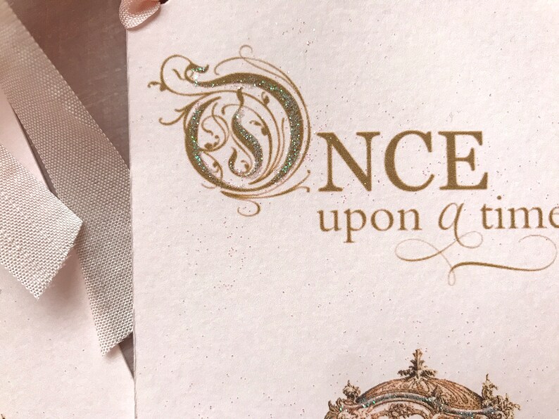 May include: A white card with gold glitter lettering that reads "ONCE upon a time". The card has a decorative gold swirl design and a pink ribbon.