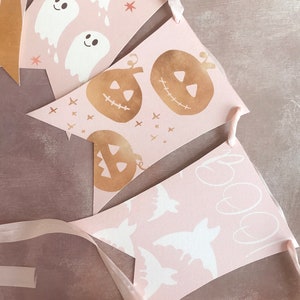 May include: A decorative bunting banner featuring triangular pennants in a soft pink hue. The pennants display white ghost illustrations, gold-toned jack-o'-lanterns, white bat silhouettes, and the handwritten text "boo!" all strung together on a ribbon.