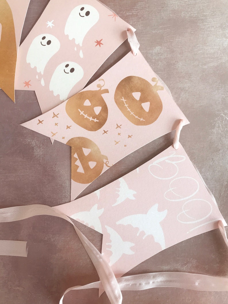 May include: A decorative bunting banner featuring triangular pennants in a soft pink hue. The pennants display white ghost illustrations, gold-toned jack-o'-lanterns, white bat silhouettes, and the handwritten text "boo!" all strung together on a ribbon.