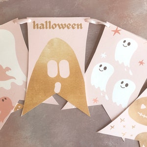 May include: A Halloween banner featuring light pink pennants strung on a ribbon. The pennants display white and gold ghost illustrations, gold pumpkin graphics, and the word halloween printed in a gold font across one of the central pennants.