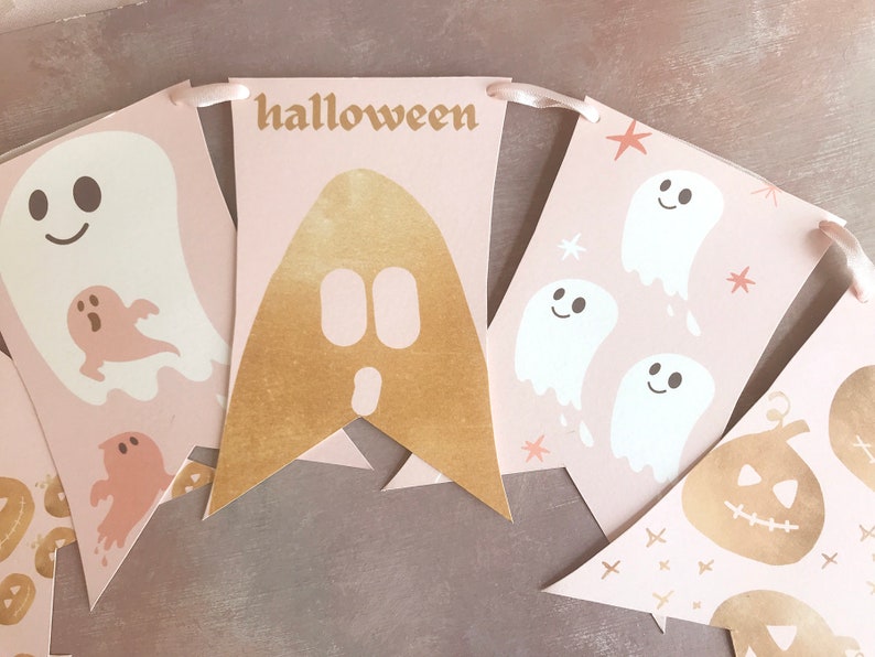 May include: A Halloween banner featuring light pink pennants strung on a ribbon. The pennants display white and gold ghost illustrations, gold pumpkin graphics, and the word halloween printed in a gold font across one of the central pennants.