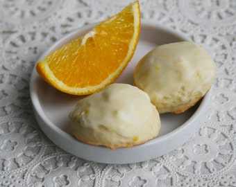 May include: Two white iced cookies with a slice of orange on a white plate. The cookies are round and have a white glaze on top. The orange slice is on the left side of the plate.