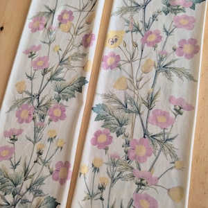 Op de afbeelding: Twee lange, rechthoekige stoffen panelen met een gebroken witte achtergrond. Elk paneel is versierd met een botanische print van roze en gele bloemen, groene bladeren en dunne stengels in een verticaal, herhalend bloemenpatroon.