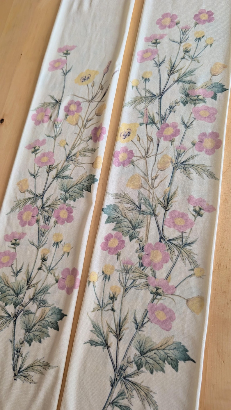 Op de afbeelding: Twee lange, rechthoekige stoffen panelen met een gebroken witte achtergrond. Elk paneel is versierd met een botanische print van roze en gele bloemen, groene bladeren en dunne stengels in een verticaal, herhalend bloemenpatroon.