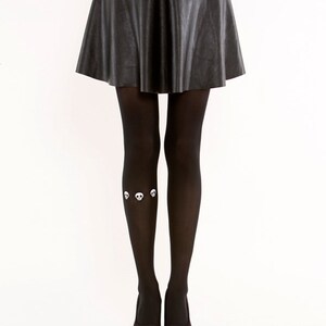 以下が含まれることがあります： 黒いフェイクレザーのスケータースカートと、右脚に3つの白いドクロのアイコンが横一列にプリントされた不透明な黒いタイツの組み合わせ。シンプルな黒いパンプスを着用しています。