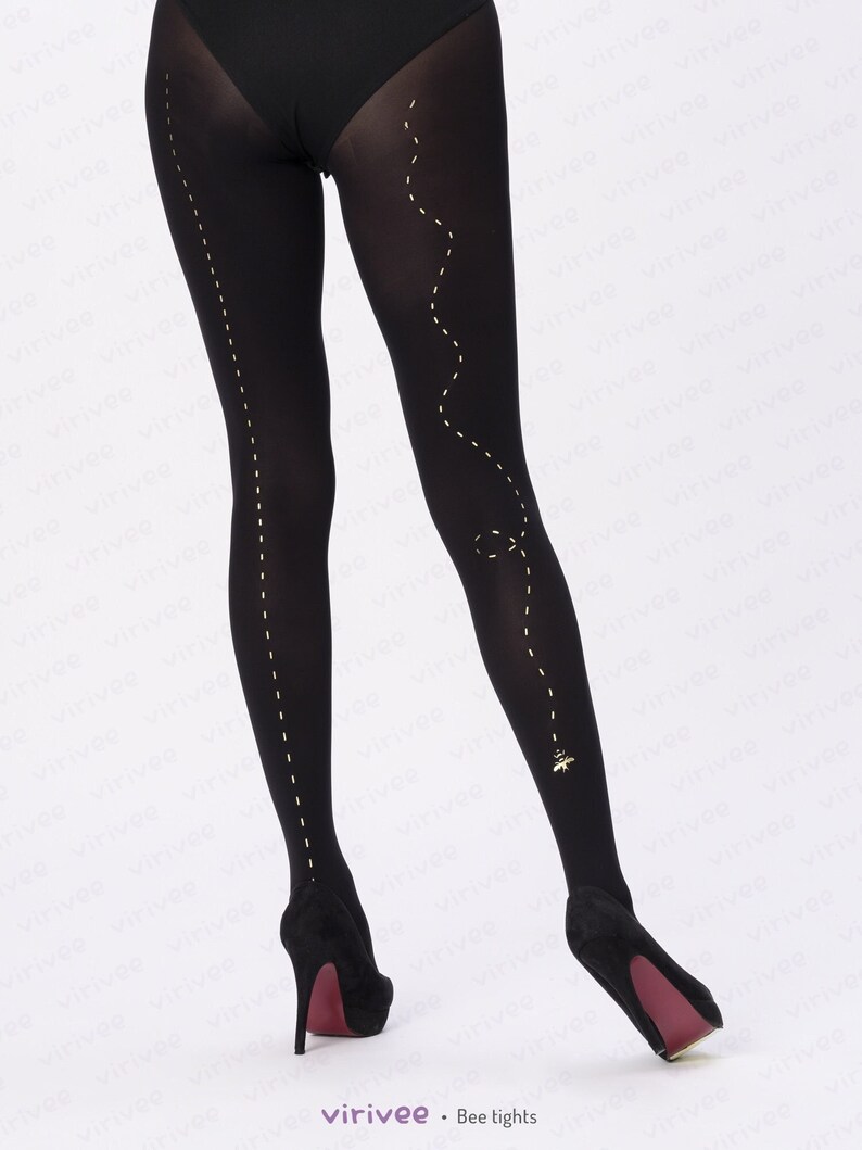May include: Opaque black tights featuring a vertical dashed line running down the outer side of each leg. The left leg includes a winding dashed path that ends with a small gold bee graphic near the ankle. The tights are paired with black heels.