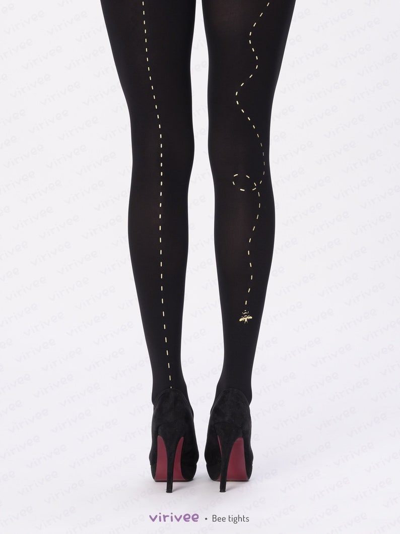 May include: Opaque black tights featuring a vertical dashed yellow line running down the center of each leg. The right leg includes a curved dashed line ending in a small yellow bee graphic near the ankle. The tights are paired with black heels.