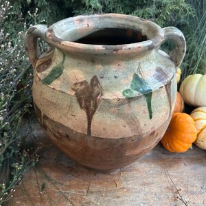 May include: A rustic, two-handled ceramic pot with a wide mouth, featuring a color palette of brown, green, and cream. The pot has a weathered appearance with dripped paint details. Small pumpkins and greenery surround the pot.