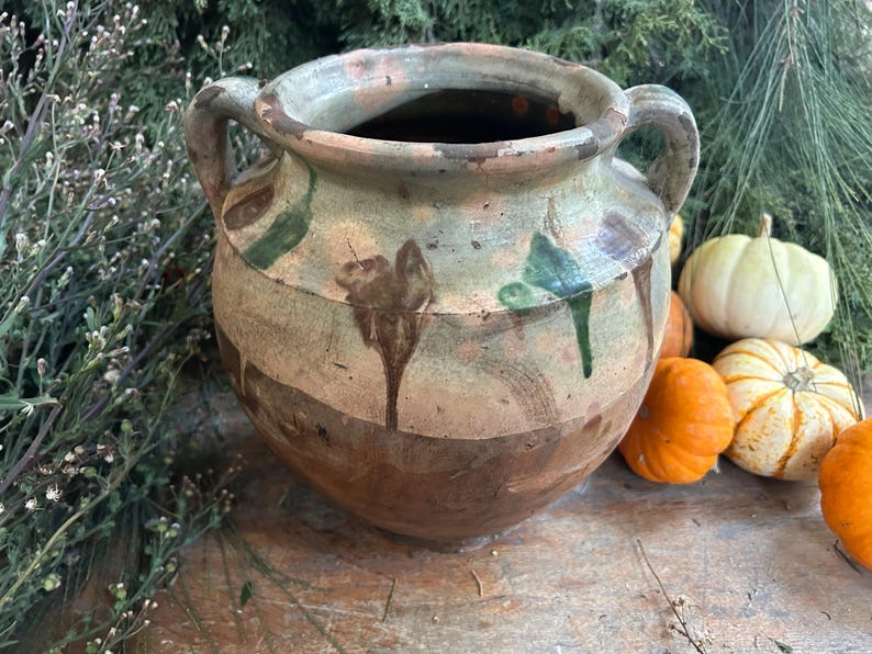 May include: A rustic, two-handled ceramic pot with a wide mouth, featuring a color palette of brown, green, and cream. The pot has a weathered appearance with dripped paint details. Small pumpkins and greenery surround the pot.