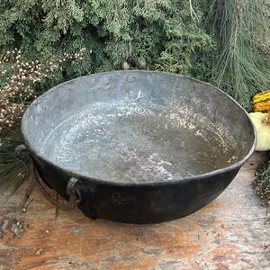 May include: A large, antique metal bowl with a dark, weathered finish. The bowl has two handles and a wide, shallow shape. The background includes greenery and several gourds, suggesting a rustic, autumnal theme. The bowl's diameter is approximately 61 cm.