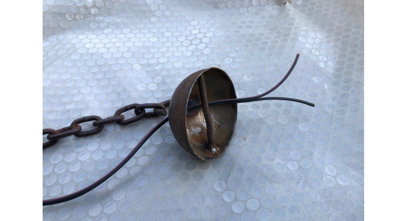K&ouml;nnte beinhalten: Eine rostige Metallkuppel mit einer Kette, die oben befestigt ist, und zwei schwarzen Dr&auml;hten, die unten herausragen.