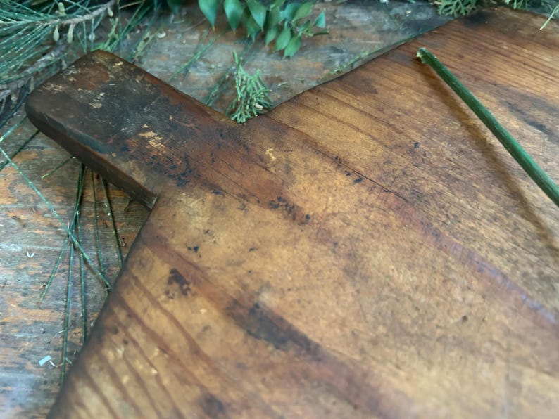 Pode incluir: Uma t&aacute;bua de corte de madeira r&uacute;stica com uma pega. A t&aacute;bua tem uma cor castanha quente com &aacute;reas mais escuras e gr&atilde;o de madeira vis&iacute;vel. Uma haste verde est&aacute; no lado direito. Para a prepara&ccedil;&atilde;o de alimentos.