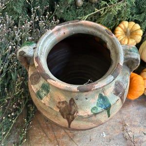 May include: A large, antique ceramic pot with a wide opening and two side handles. The pot has a weathered appearance with brown, green, and blue painted details. Small pumpkins in various colors and shapes are arranged around the pot, suggesting a fall theme.