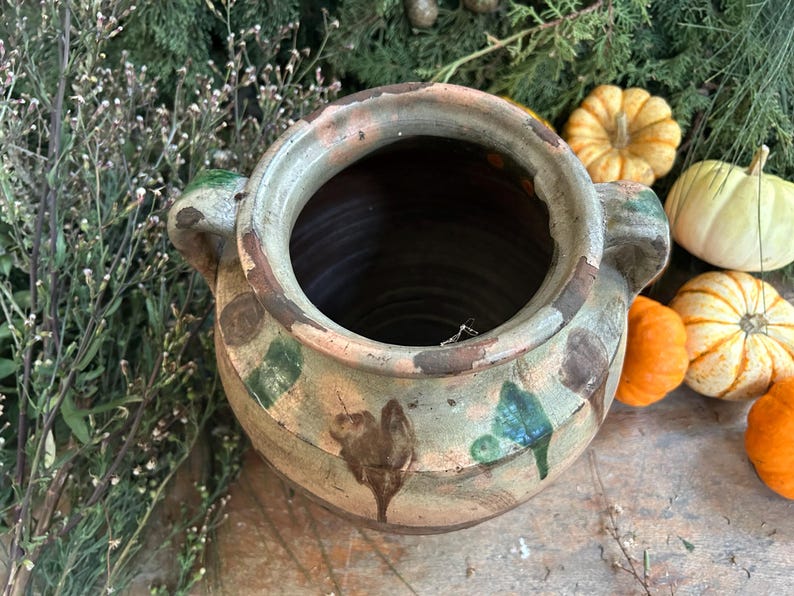 May include: A large, antique ceramic pot with a wide opening and two side handles. The pot has a weathered appearance with brown, green, and blue painted details. Small pumpkins in various colors and shapes are arranged around the pot, suggesting a fall theme.