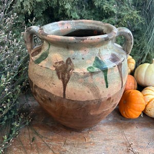 May include: A rustic, two-handled ceramic pot with a wide mouth, featuring a brown and tan color scheme with green and brown accents. The pot is surrounded by small pumpkins and greenery, suggesting a fall theme.