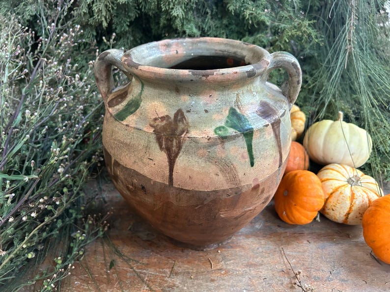 May include: A rustic, two-handled ceramic pot with a wide mouth, featuring a brown and tan color scheme with green and brown accents. The pot is surrounded by small pumpkins and greenery, suggesting a fall theme.