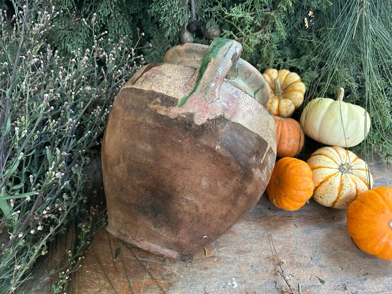 May include: A rustic, brown ceramic jug with a green handle, surrounded by small pumpkins in shades of orange, yellow, and white. The arrangement is set against a backdrop of greenery, creating a fall harvest theme.