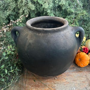Puede incluir: Una olla grande de cerámica gris oscuro con dos asas. La olla es redonda con una abertura ancha. El fondo incluye vegetación y pequeñas calabazas, lo que sugiere un tema otoñal.
