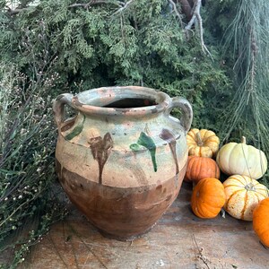 May include: A rustic, two-handled ceramic pot with a brown base and a light green and brown upper section. The pot has a weathered appearance with drips of green glaze. Miniature pumpkins in various colors are arranged nearby.