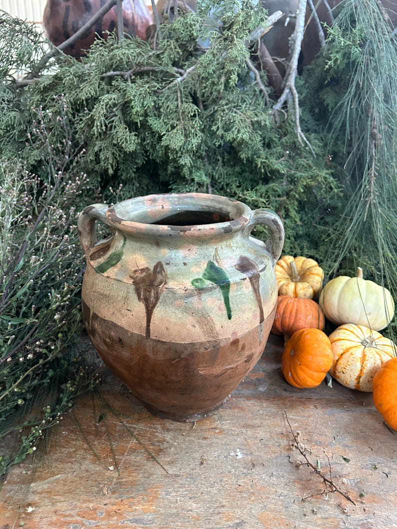 May include: A rustic, two-handled ceramic pot with a brown base and a light green and brown upper section. The pot has a weathered appearance with drips of green glaze. Miniature pumpkins in various colors are arranged nearby.
