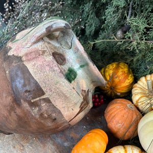 May include: A rustic, hand-painted ceramic pot with a handle, featuring brown, beige, and green accents. The pot is surrounded by small pumpkins in shades of orange, yellow, and white, with red berries and green foliage.
