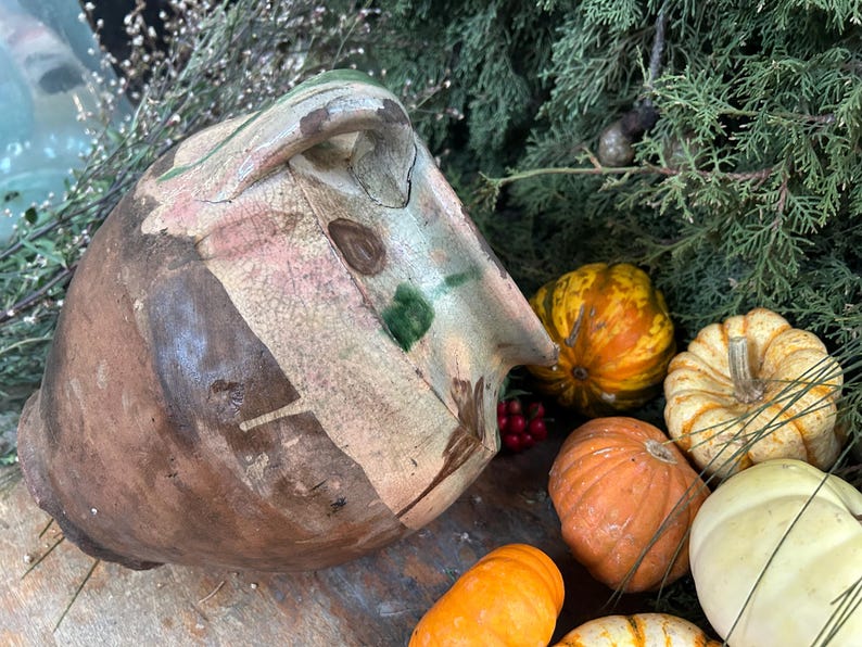 May include: A rustic, hand-painted ceramic pot with a handle, featuring brown, beige, and green accents. The pot is surrounded by small pumpkins in shades of orange, yellow, and white, with red berries and green foliage.