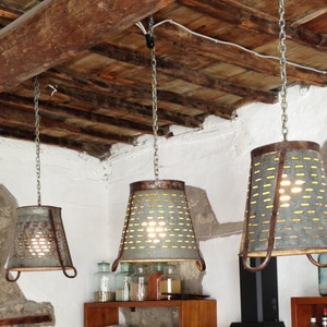 May include: Three rustic metal pendant lights hang from a wooden ceiling. Each light is made from a repurposed metal container with a perforated design. The lights are illuminated with warm white bulbs, casting a soft glow.