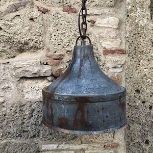 Puede incluir: Una lámpara colgante de metal rústico con una pantalla en forma de campana, colgada de una cadena contra una pared de piedra.
