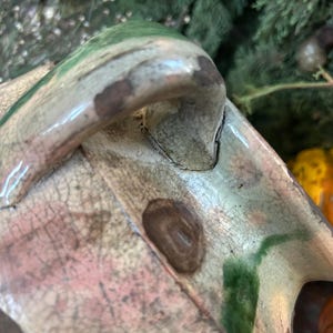 May include: Close-up of a ceramic object with a handle, featuring a blend of colors including green, pink, and brown. The surface has a cracked, aged appearance. The background includes greenery and small gourds.
