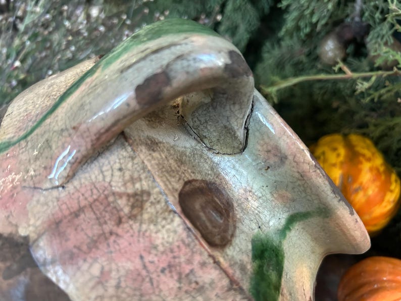 May include: Close-up of a ceramic object with a handle, featuring a blend of colors including green, pink, and brown. The surface has a cracked, aged appearance. The background includes greenery and small gourds.