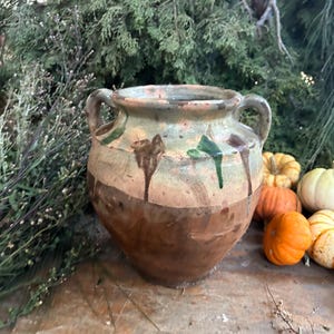 May include: A rustic, two-handled ceramic pot with a wide base and a narrow neck. The pot features a brown, tan, and green color scheme with dripped paint details. Small pumpkins and greenery are in the background.