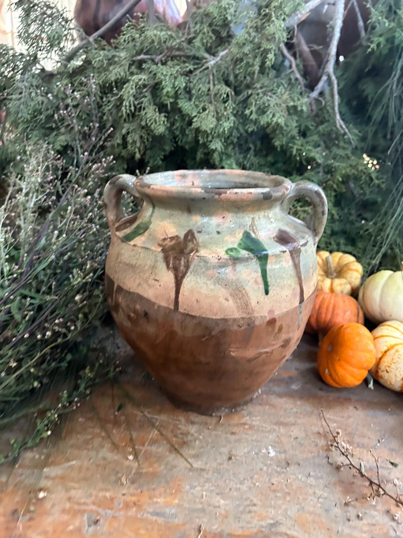 May include: A rustic, two-handled ceramic pot with a wide base and a narrow neck. The pot features a brown, tan, and green color scheme with dripped paint details. Small pumpkins and greenery are in the background.