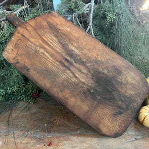 Pode incluir: Uma t&aacute;bua de corte retangular de madeira com uma al&ccedil;a, em um tom marrom quente com &aacute;reas mais escuras. A t&aacute;bua mostra sinais de uso. Ab&oacute;boras decorativas e vegeta&ccedil;&atilde;o circundam a t&aacute;bua, sugerindo um tema de outono.