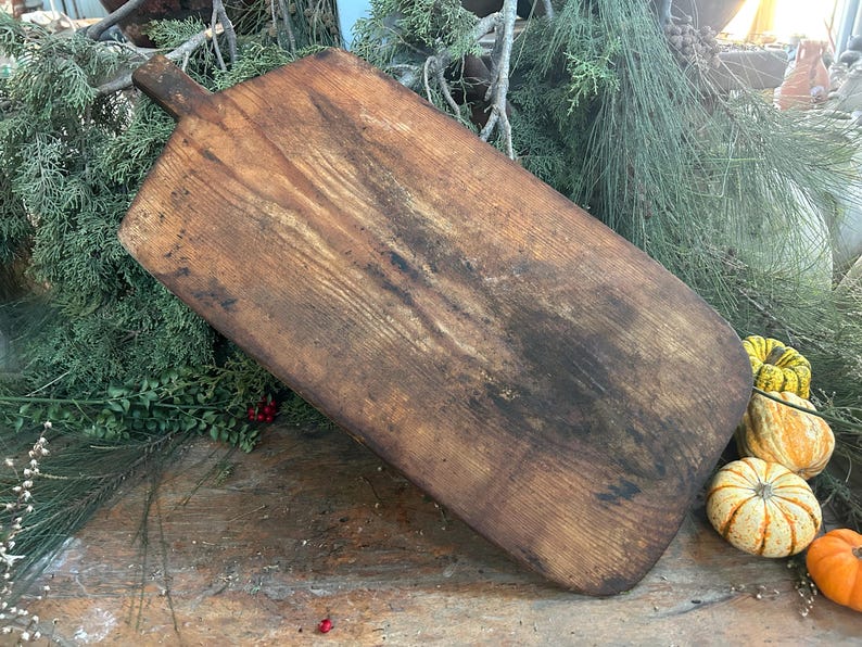 Pode incluir: Uma t&aacute;bua de corte retangular de madeira com uma al&ccedil;a, em um tom marrom quente com &aacute;reas mais escuras. A t&aacute;bua mostra sinais de uso. Ab&oacute;boras decorativas e vegeta&ccedil;&atilde;o circundam a t&aacute;bua, sugerindo um tema de outono.