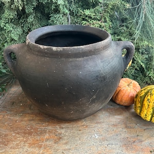 Puede incluir: Una olla grande de cerámica rústica de color gris oscuro con dos asas. La olla se encuentra sobre una superficie de madera con pequeñas calabazas y vegetación en el fondo. La olla tiene una abertura ancha y un cuerpo redondeado.
