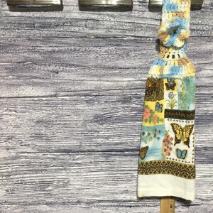 Peut inclure: Un torchon de cuisine suspendu avec un dessus crochet&eacute; bleu, jaune et beige. Le corps du torchon pr&eacute;sente un motif patchwork avec des papillons et des motifs floraux de diff&eacute;rentes couleurs, dont le marron, le jaune et le bleu. Le bord inf&eacute;rieur est blanc.
