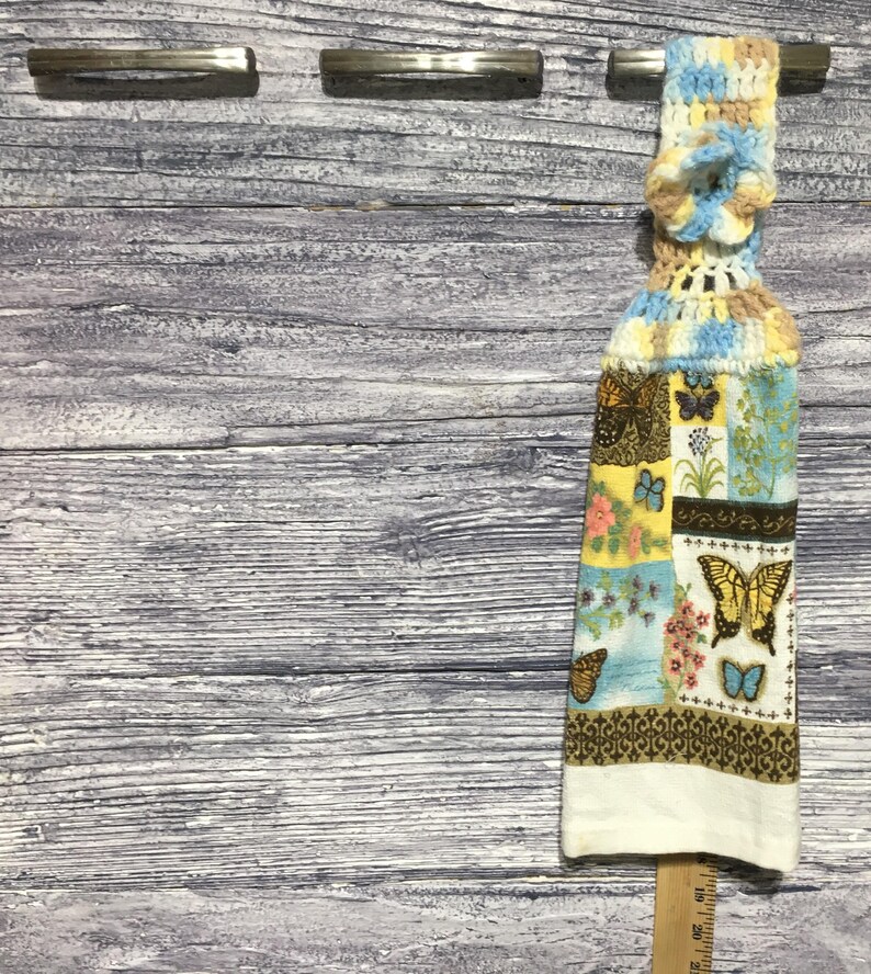 Peut inclure: Un torchon de cuisine suspendu avec un dessus crochet&eacute; bleu, jaune et beige. Le corps du torchon pr&eacute;sente un motif patchwork avec des papillons et des motifs floraux de diff&eacute;rentes couleurs, dont le marron, le jaune et le bleu. Le bord inf&eacute;rieur est blanc.