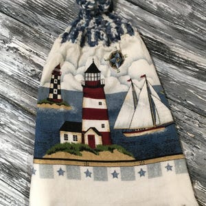 May include: A nautical kitchen towel with a hanging loop. The design includes a red and white striped lighthouse, a black and white lighthouse, a sailboat, and a compass. The bottom edge has a fringe and blue star accents.