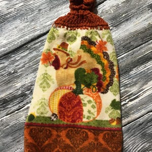 May include: A fall-themed kitchen towel with a crochet top. The towel features a print of pumpkins, leaves, and a turkey. The bottom border has a repeating heart pattern in shades of orange and brown.