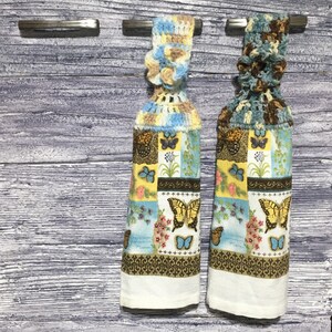 Peut inclure: Deux essuie-mains de cuisine d&eacute;coratifs. Chaque serviette pr&eacute;sente un motif patchwork avec des papillons, des fleurs et des motifs g&eacute;om&eacute;triques en jaune, bleu et marron. Les hauts sont crochet&eacute;s dans des couleurs assorties, et les bas sont blancs.