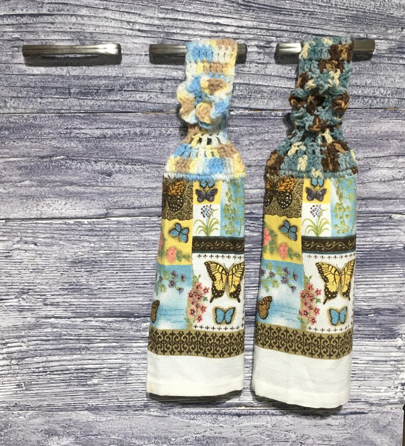 Peut inclure: Deux essuie-mains de cuisine d&eacute;coratifs. Chaque serviette pr&eacute;sente un motif patchwork avec des papillons, des fleurs et des motifs g&eacute;om&eacute;triques en jaune, bleu et marron. Les hauts sont crochet&eacute;s dans des couleurs assorties, et les bas sont blancs.