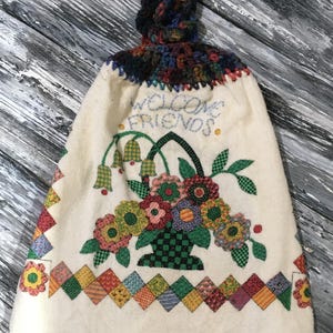 Op de afbeelding: Crèmekleurige keukentowel met een gehaakte bovenkant en een bloemenmandontwerp. De handdoek heeft de woorden "WELCOME FRIENDS" geborduurd boven de mand. De onderkant heeft een patchwork-ontwerp met kleurrijke vierkanten.