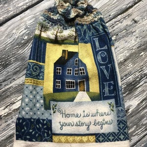 May include: A kitchen towel featuring a blue house design, the word "LOVE", and the phrase "Home is where your story begins". The towel has a crocheted top and is made of fabric with various patterns and colours, suitable for the kitchen.