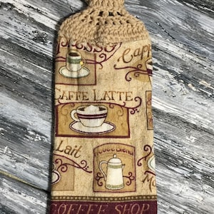 May include: A beige crocheted kitchen towel holder with a button closure. The towel features coffee-themed graphics, including cups, a coffee pot, and the words "Caffe Latte" and "Coffee Shop."