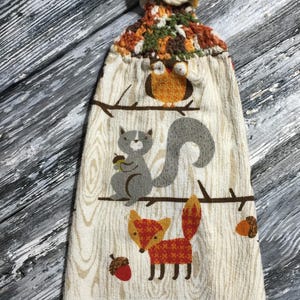 May include: A hanging kitchen towel with a crochet top and button fastening. The towel features a woodland scene with an owl, squirrel, and fox on a wood-grain background. The crochet top is made of brown, orange, and cream yarn.